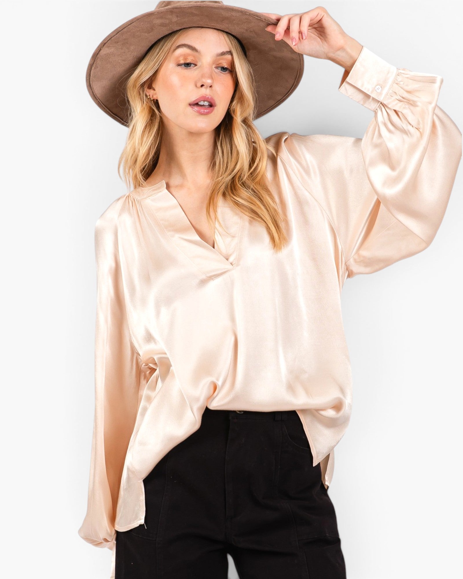 A woman wearing a light beige satin blouse with long balloon sleeves and a V-neckline, paired with black pants and a brown wide-brim hat, against a plain white background.