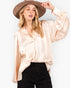 A woman wearing a light beige satin blouse with long balloon sleeves and a V-neckline, paired with black pants and a brown wide-brim hat, against a plain white background.