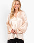 A woman wearing a shiny champagne beige satin blouse with a split V-neckline and voluminous long sleeves, paired with black pants, against a plain white background.