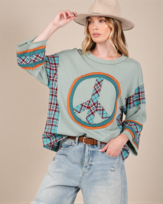 Woman wearing a light green sweatshirt with a peace symbol design, plaid sleeves, and a beige hat on a beige background.