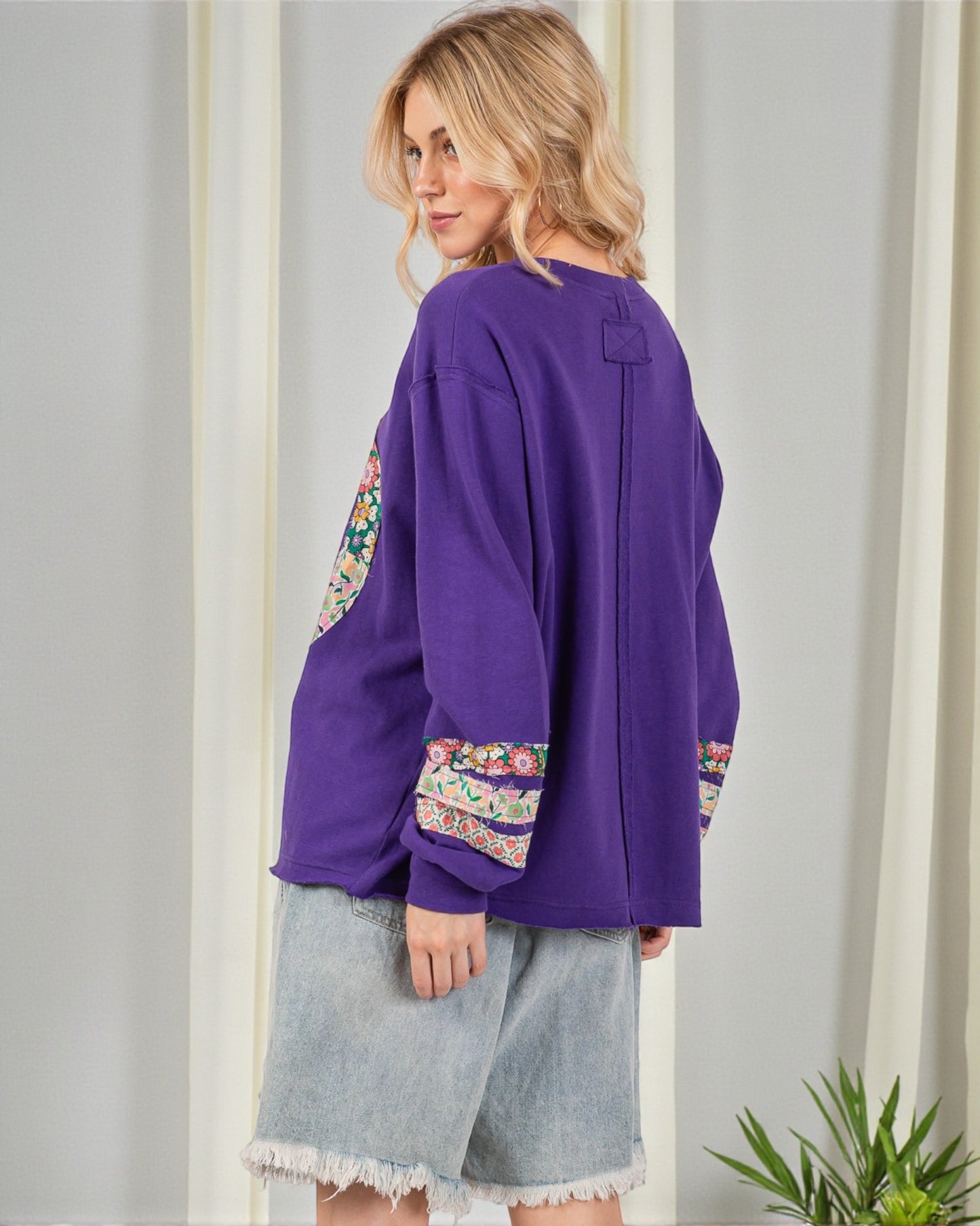 Woman wearing a purple top with floral cuffs and hem, standing against a white wall.