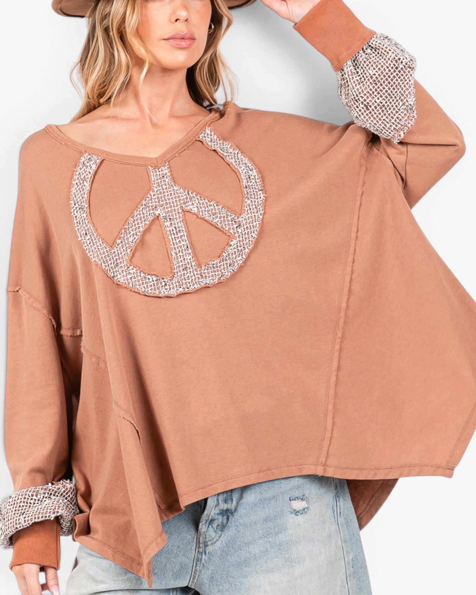 A woman wearing a loose, oversized long-sleeve top in a warm taupe-brown color, featuring a large peace sign motif on the chest and textured beaded cuffs, paired with light-wash jeans.