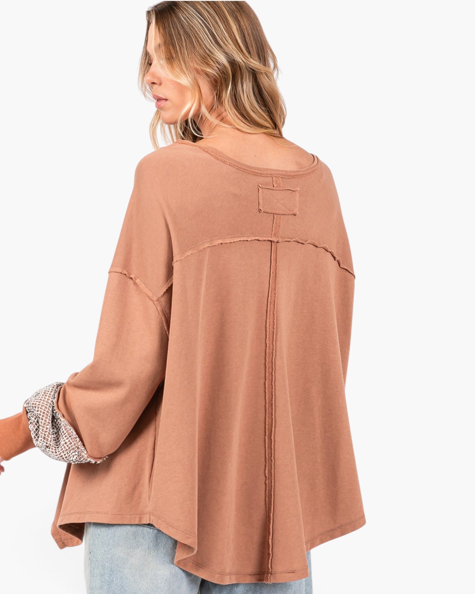 A woman shown from the back wearing a loose, dusty rose-brown top with three-quarter sleeves, a small back patch detail, and a patterned cuff peeking out from one sleeve, paired with light blue jeans against a plain white background.