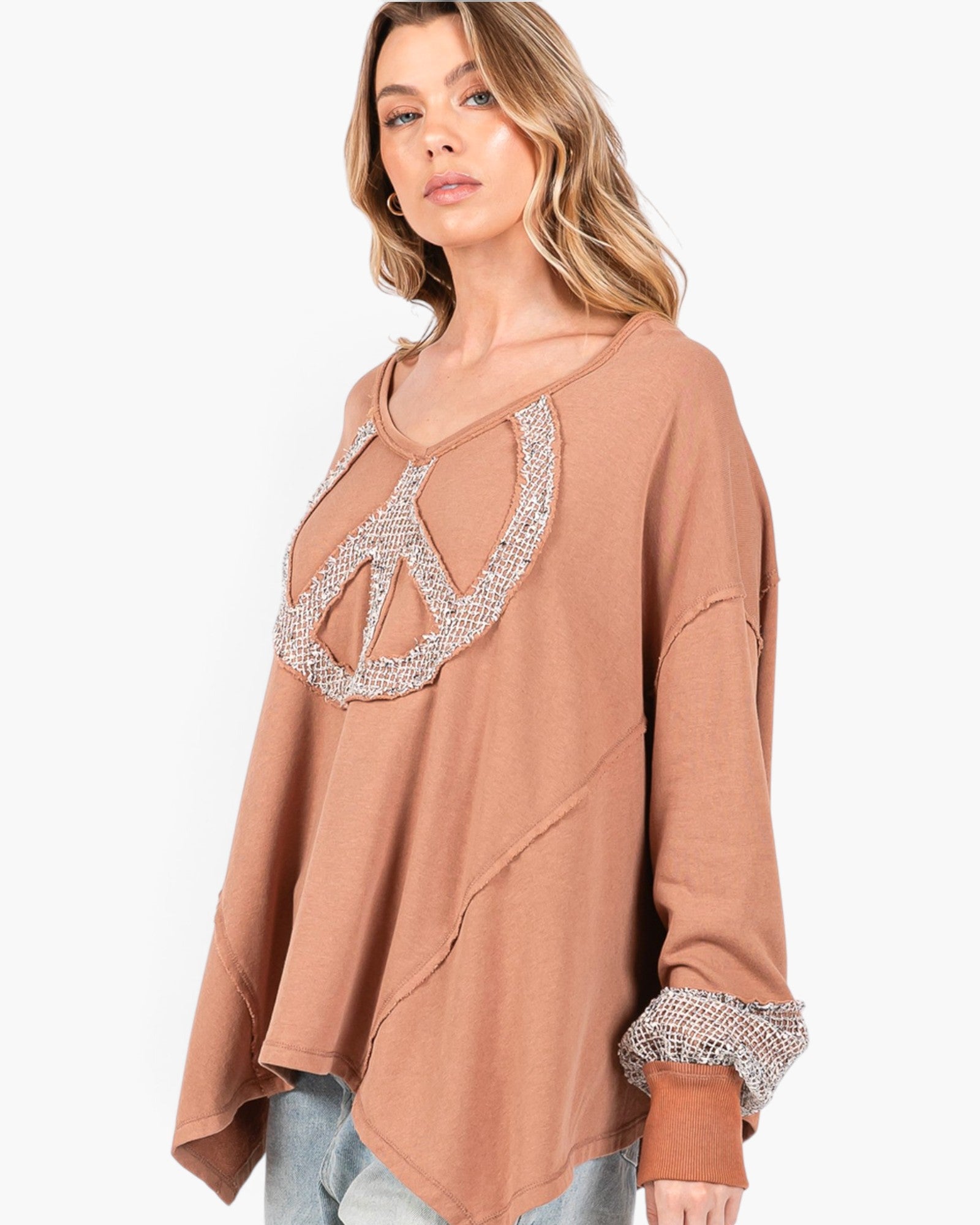 A woman wearing a loose, long-sleeve taupe-brown top with a peace sign cutout embellished with silver rhinestones, paired with light blue jeans, against a plain white background.