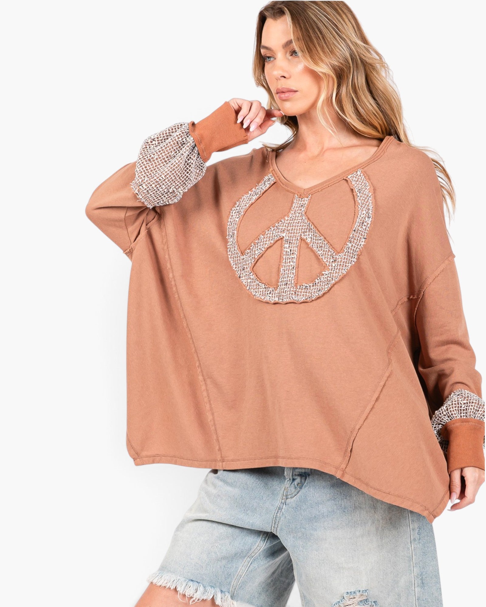 A woman wearing a loose, oversized tan long-sleeve top with a large rhinestone-embellished peace sign on the chest and matching rhinestone cuffs, paired with light-wash distressed denim shorts against a plain white background.