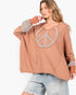 A woman wearing a loose, oversized tan long-sleeve top with a large rhinestone-embellished peace sign on the chest and matching rhinestone cuffs, paired with light-wash distressed denim shorts against a plain white background.