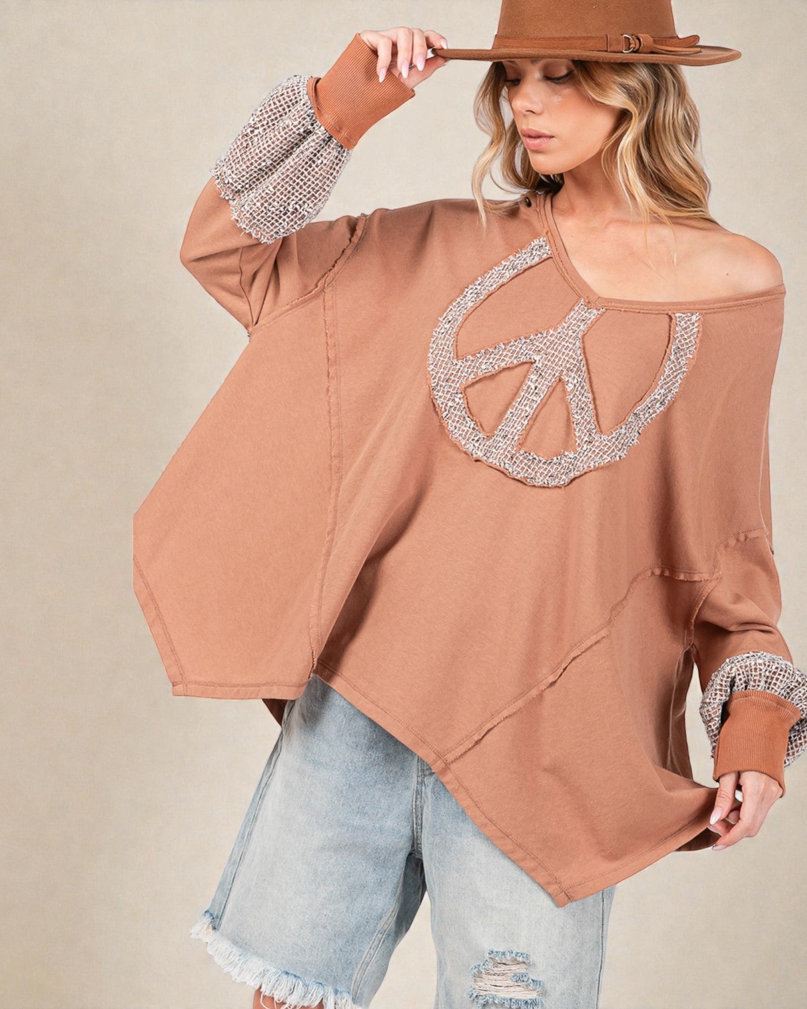 Woman wearing a brown off-shoulder top with a peace sign design and rhinestone details, holding a brown hat.