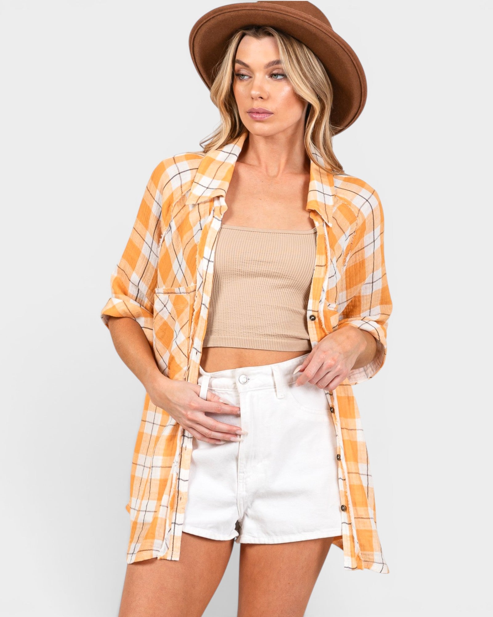 Woman wearing a yellow plaid shirt, beige top, white shorts, and brown hat on a light gray background