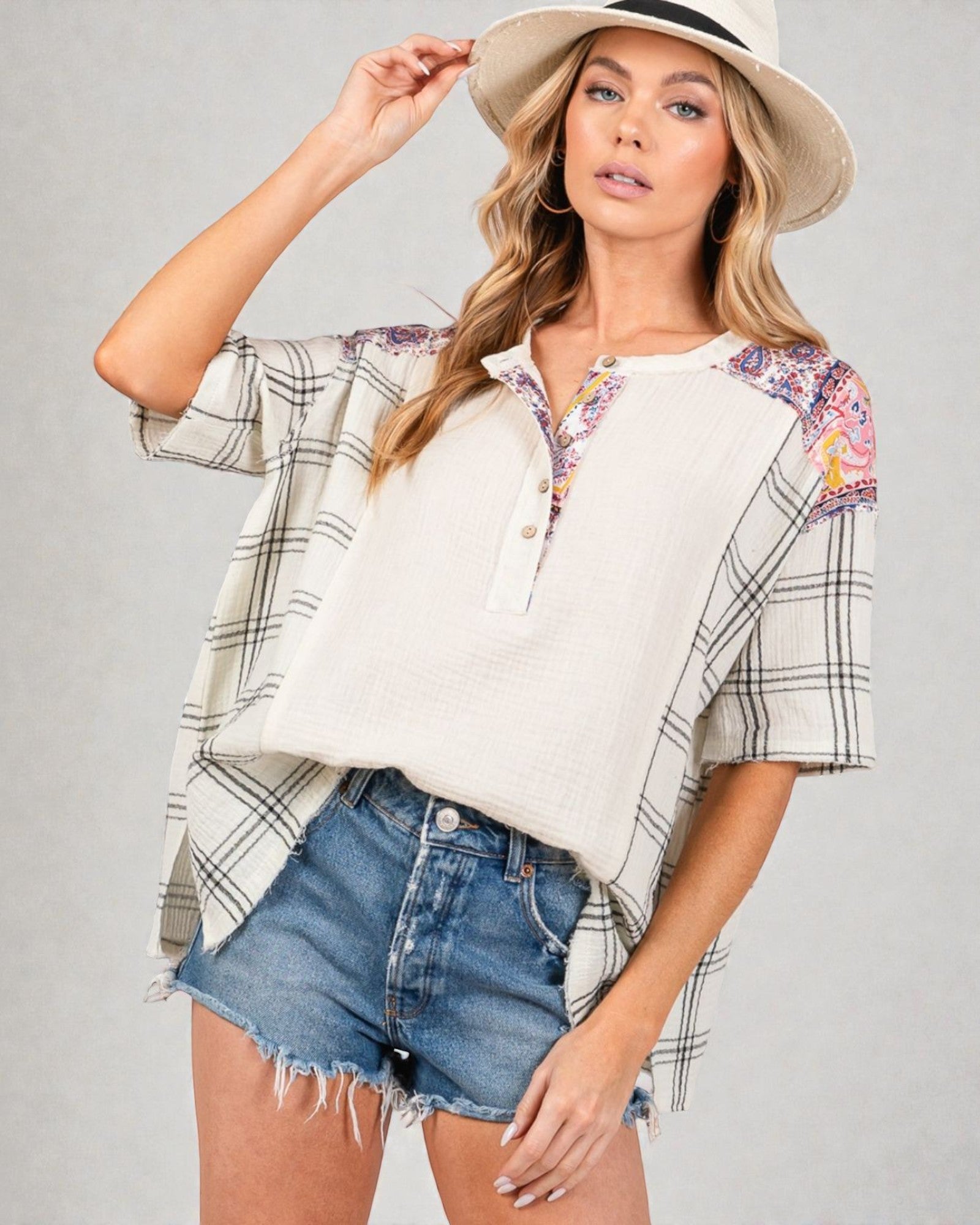 Woman wearing a plaid shirt over a floral top with denim shorts and a hat on a gray background