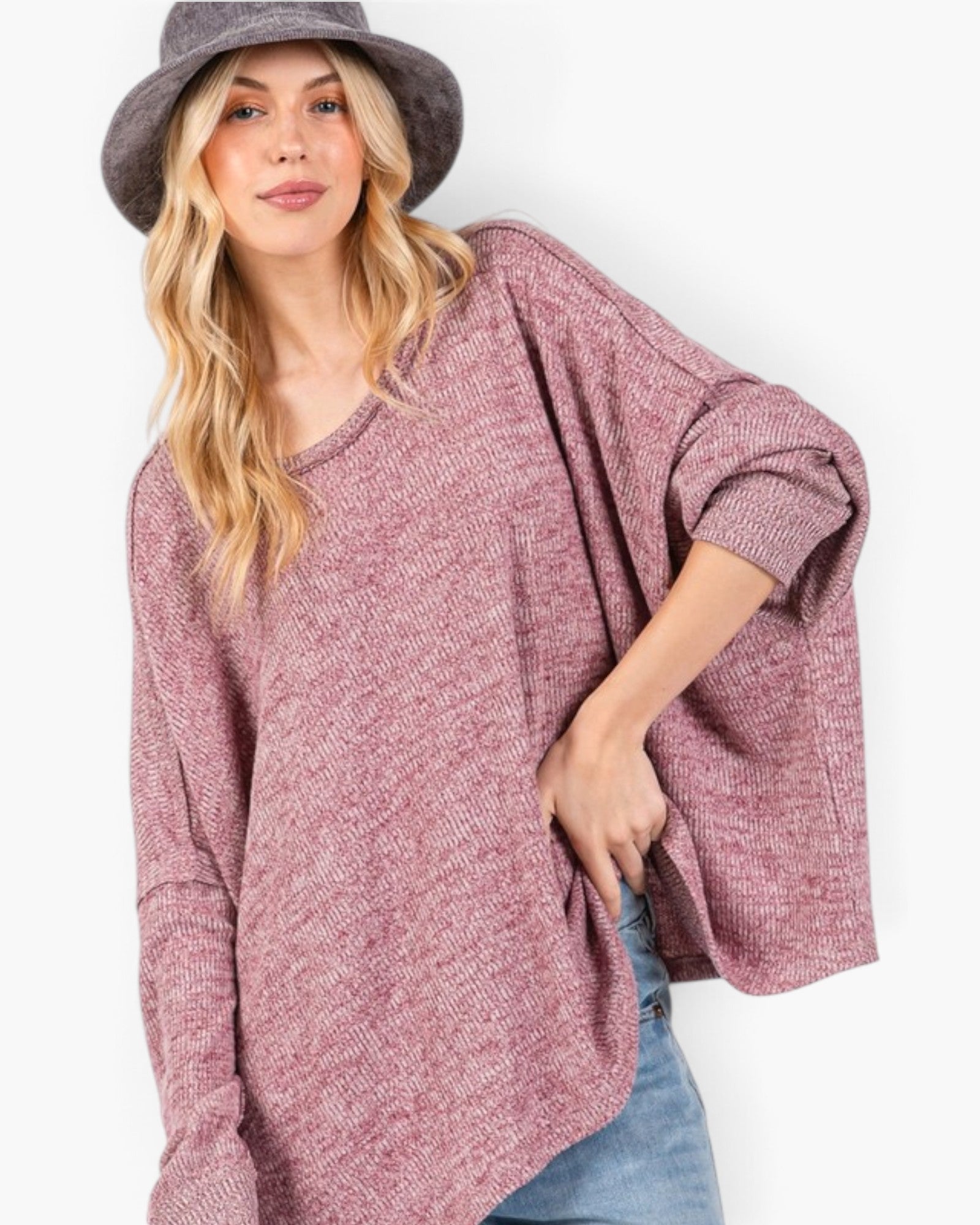 A woman wearing a loose, heathered dusty rose knit sweater with wide three-quarter sleeves, paired with blue jeans and a gray bucket hat, against a plain white background.