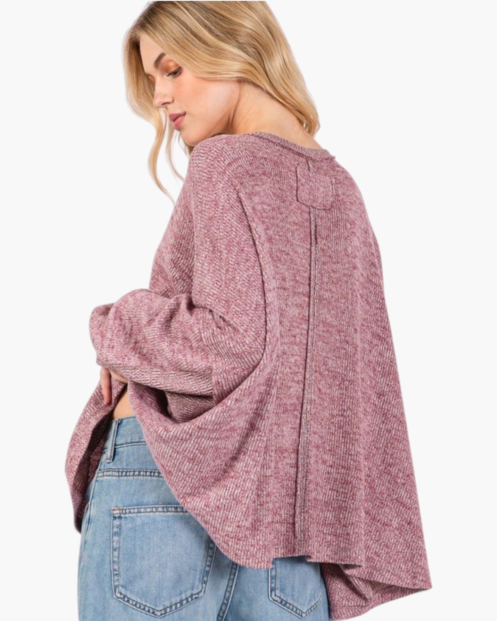 A woman wearing a dusty rose heathered knit sweater with wide bell sleeves and a relaxed fit, paired with blue jeans, shown from a side-back angle against a plain white background.
