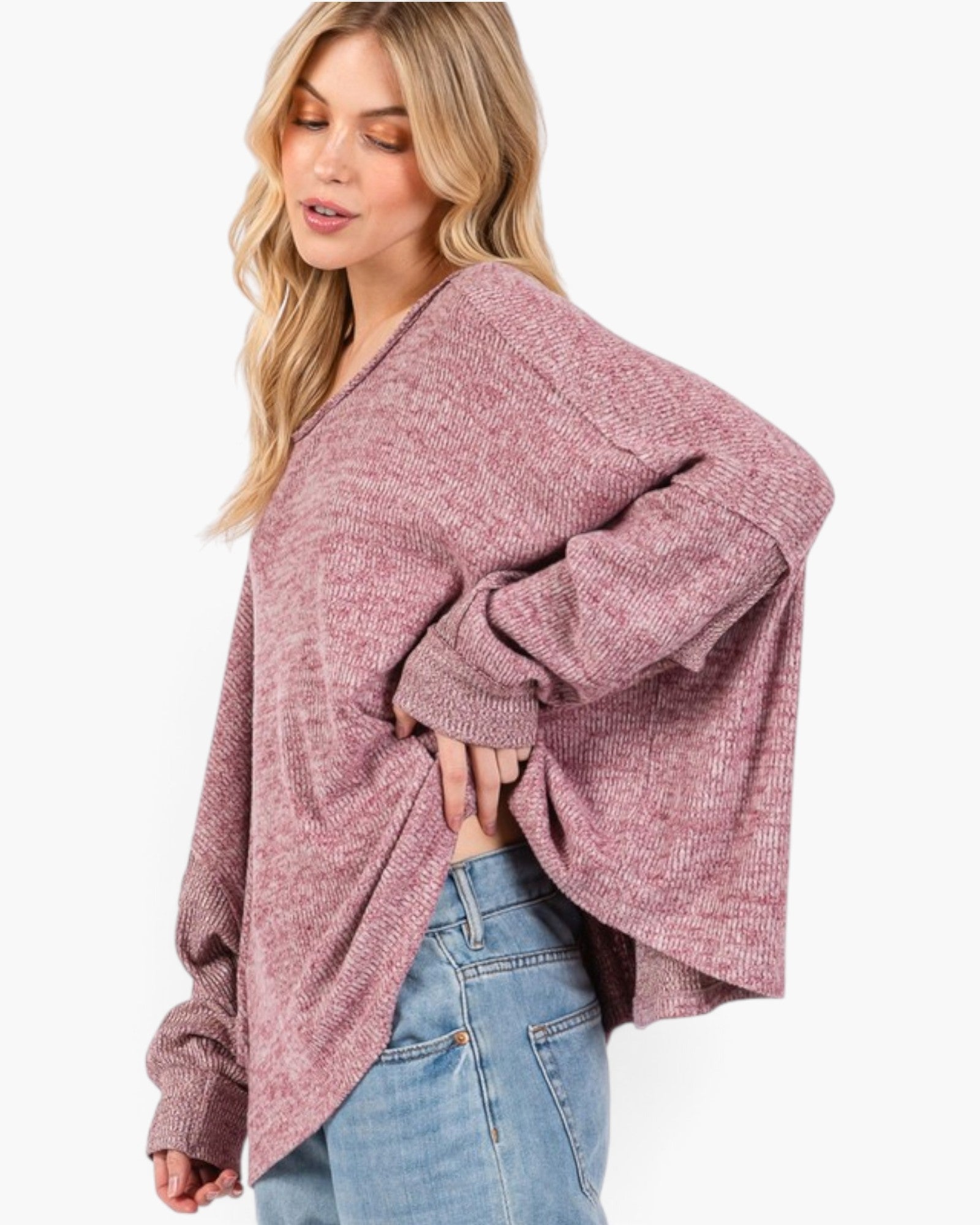 A woman wearing a loose, heathered dusty rose knit sweater with wide sleeves and light blue jeans, photographed against a clean white background.