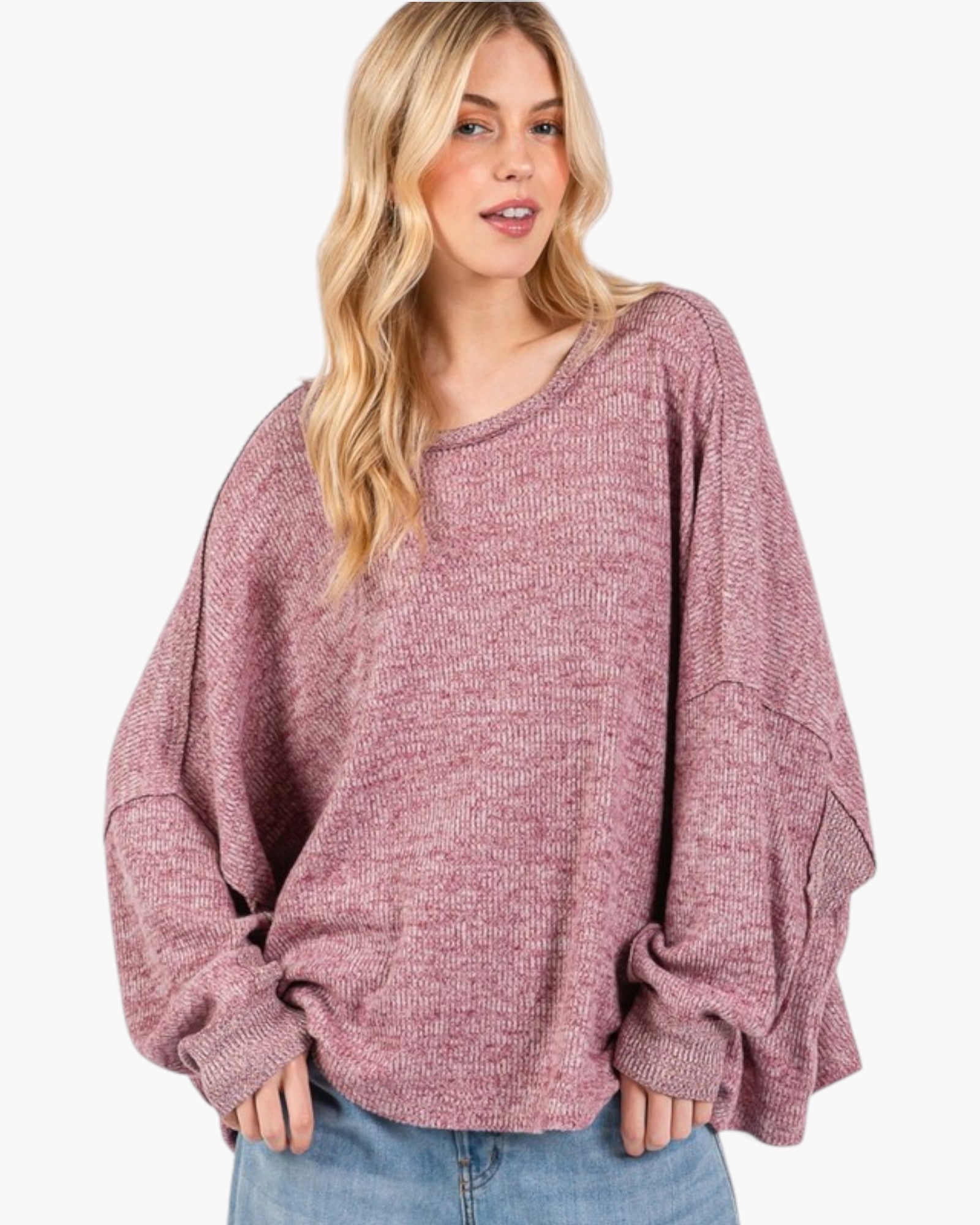 A woman wearing a loose, oversized knit sweater in a heathered dusty rose color, paired with blue jeans, against a plain white background.