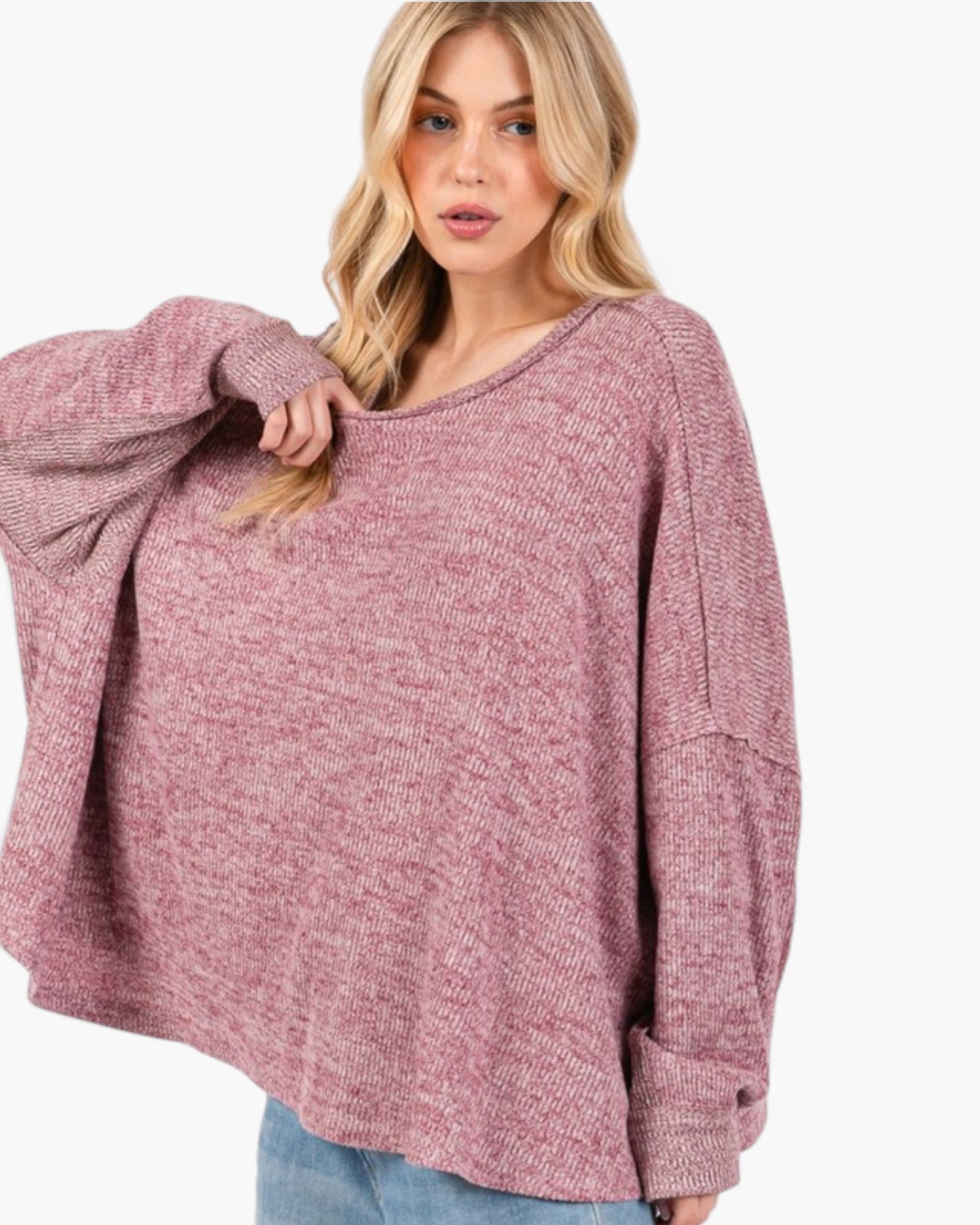 A woman wearing a loose, oversized knit sweater in a heathered dusty rose color, paired with light blue jeans, against a plain white background.