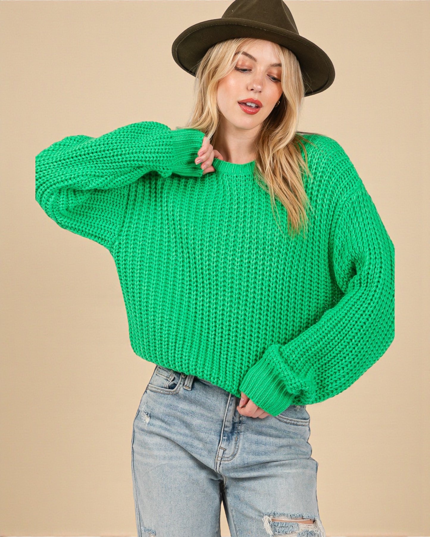 Person wearing a bright green chunky knit sweater and a brown hat on a beige background
