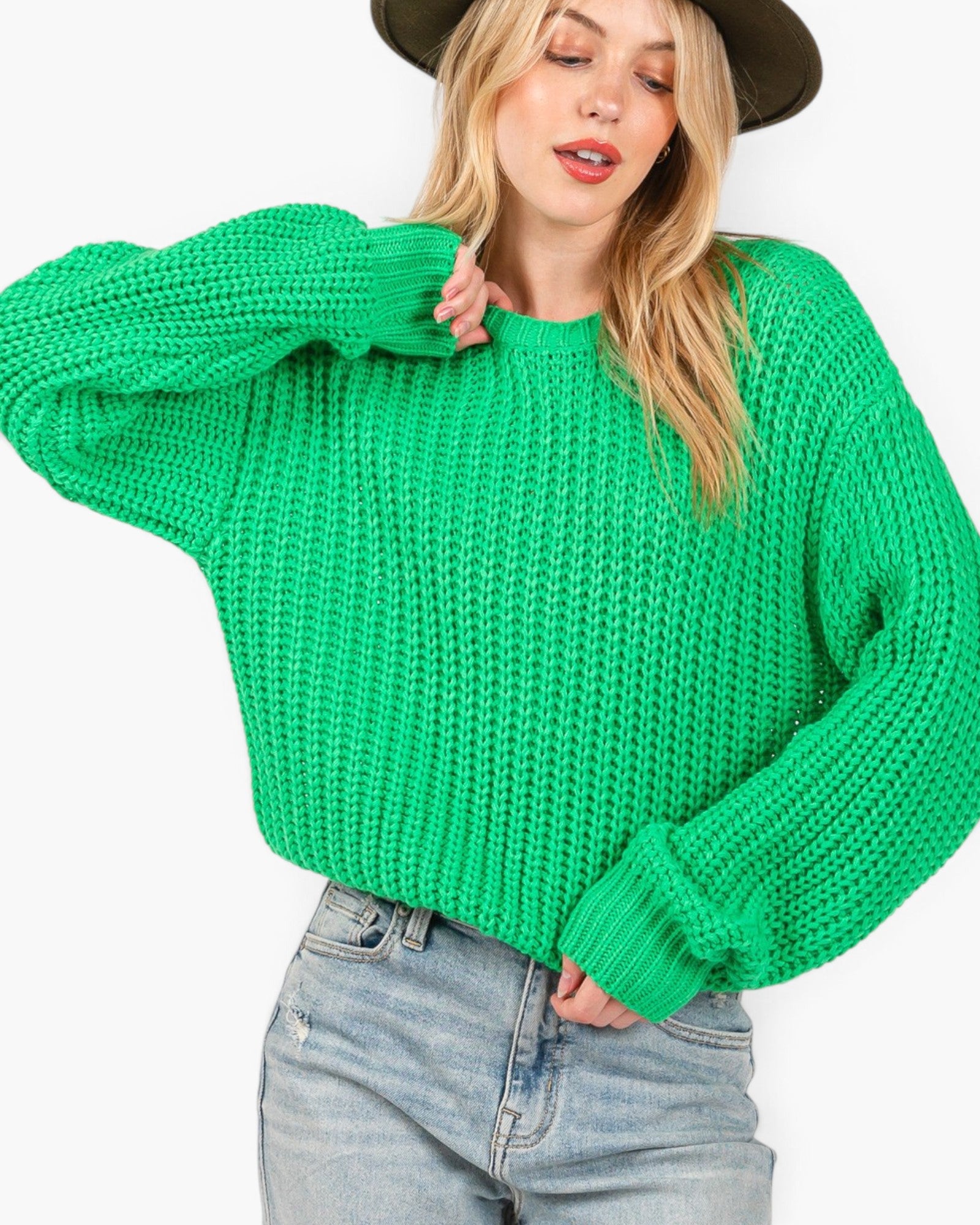A woman wearing a bright green chunky knit sweater and light-wash blue jeans, cropped from the hat brim down to the hips, against a plain white background.