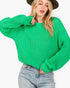 A woman wearing a bright green chunky knit sweater and light-wash blue jeans, cropped from the hat brim down to the hips, against a plain white background.