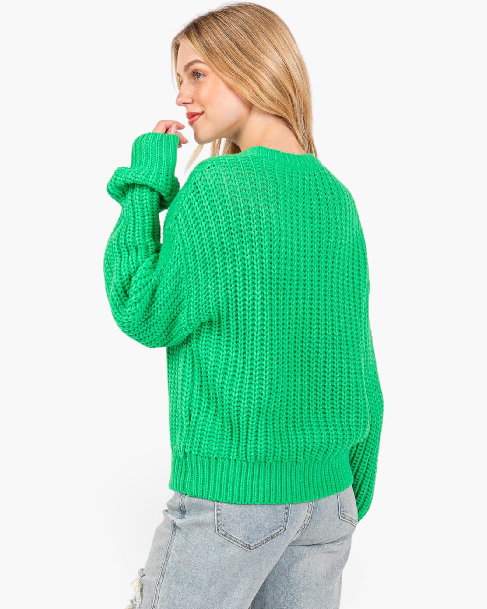 A woman wearing a bright green chunky knit sweater and light wash jeans, shown from a side-back angle against a plain white background.