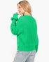 A woman wearing a bright green chunky knit sweater and light wash jeans, shown from a side-back angle against a plain white background.