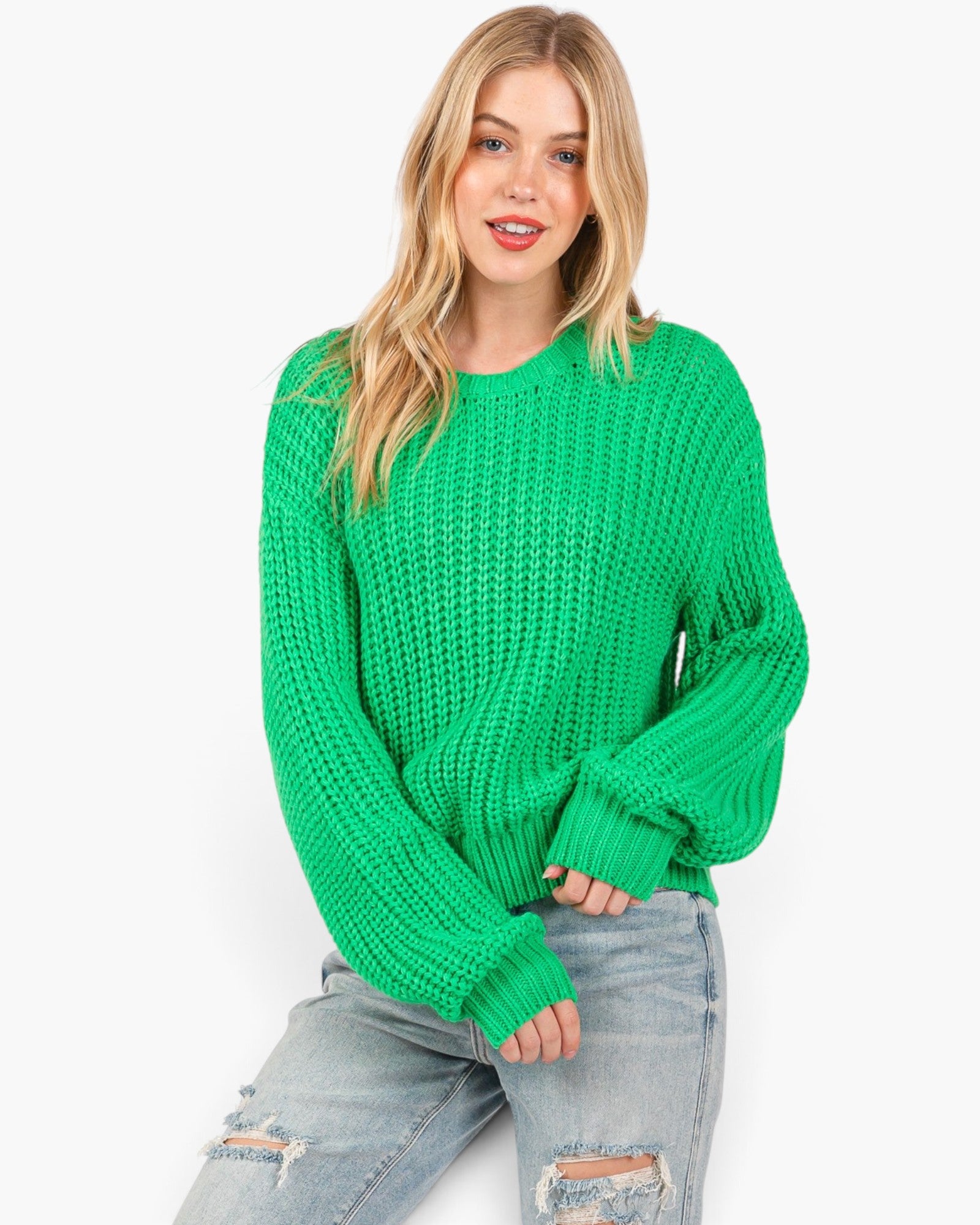 A woman wearing a bright green chunky knit sweater and light-wash distressed jeans, posed against a clean white studio background.