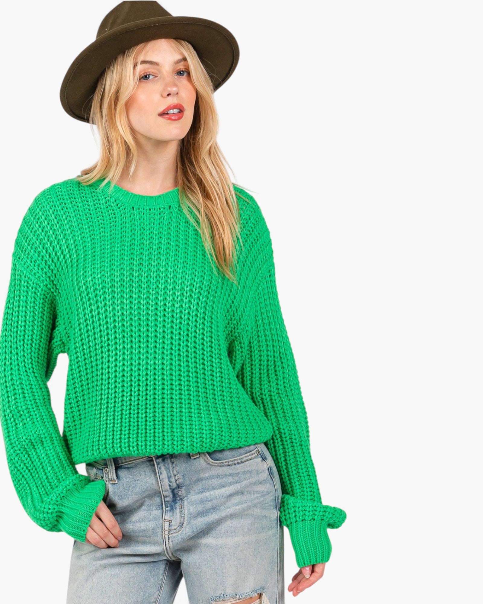A woman wearing a bright green chunky knit sweater, light-wash distressed jeans, and a brown wide-brim hat, photographed against a plain light gray studio background.