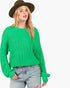 A woman wearing a bright green chunky knit sweater, light-wash distressed jeans, and a brown wide-brim hat, photographed against a plain light gray studio background.