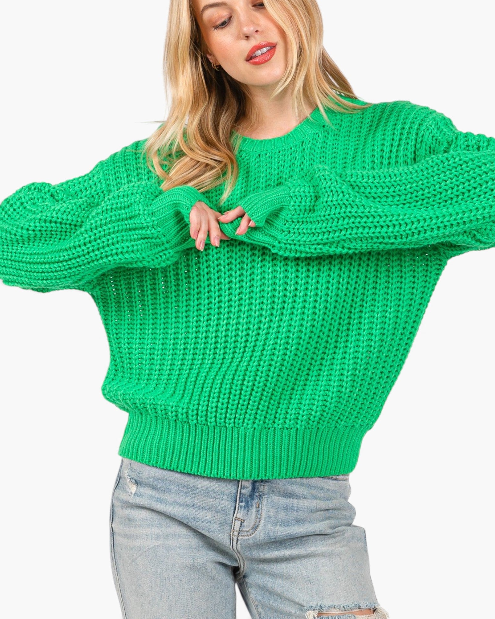 A woman wearing a bright green chunky knit sweater with oversized sleeves and light-wash distressed jeans, shown against a plain light gray background.