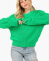A woman wearing a bright green chunky knit sweater with oversized sleeves and light-wash distressed jeans, shown against a plain light gray background.