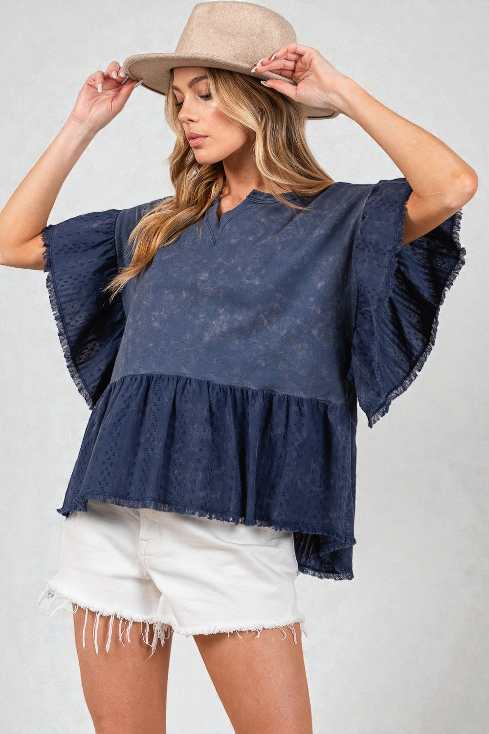 Woman wearing a navy blue top with ruffled sleeves and white shorts, holding a beige hat.