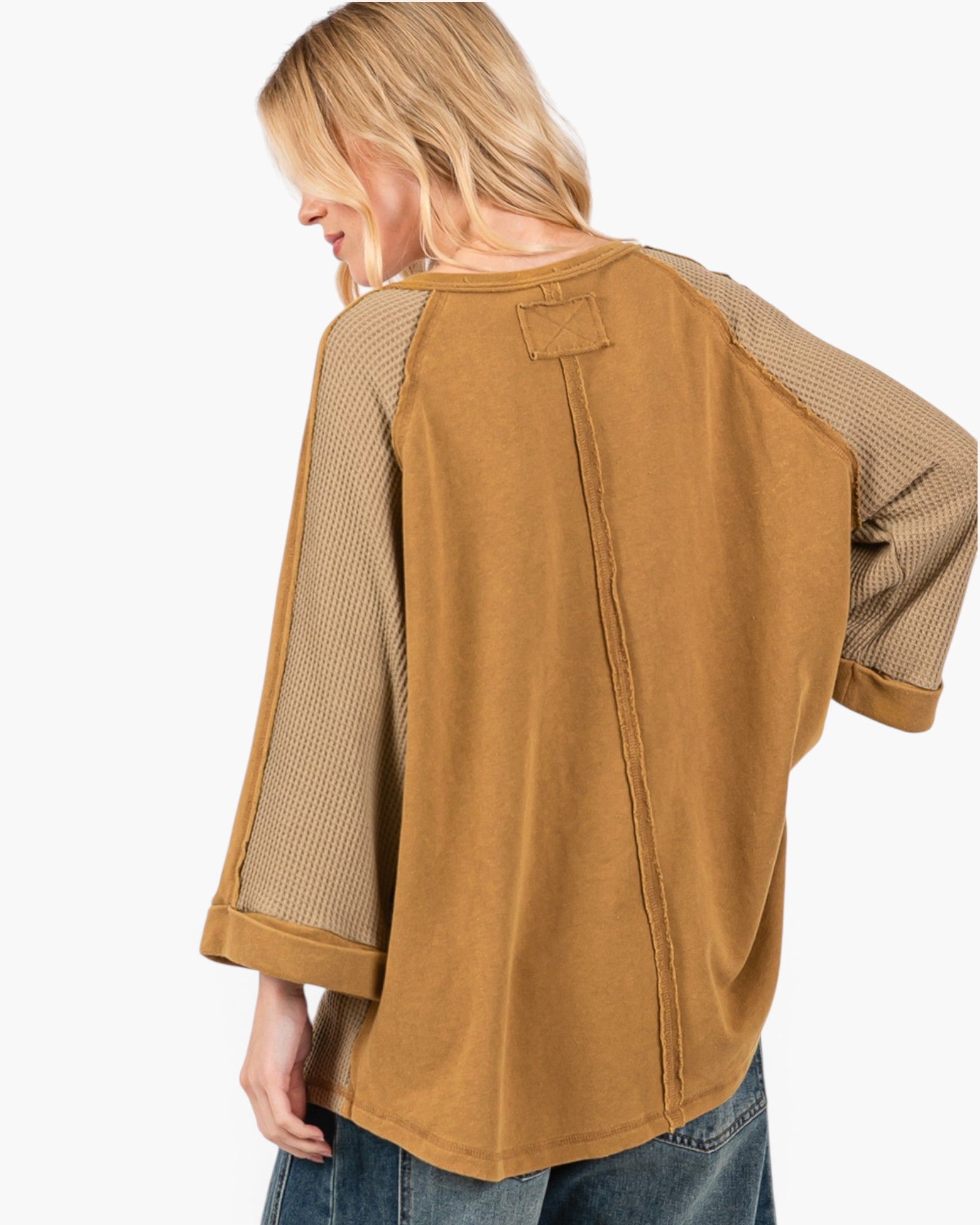 A woman shown from the back wearing a tan and beige long-sleeve top with textured mesh sleeves and a back seam detail, paired with blue jeans against a plain white background.