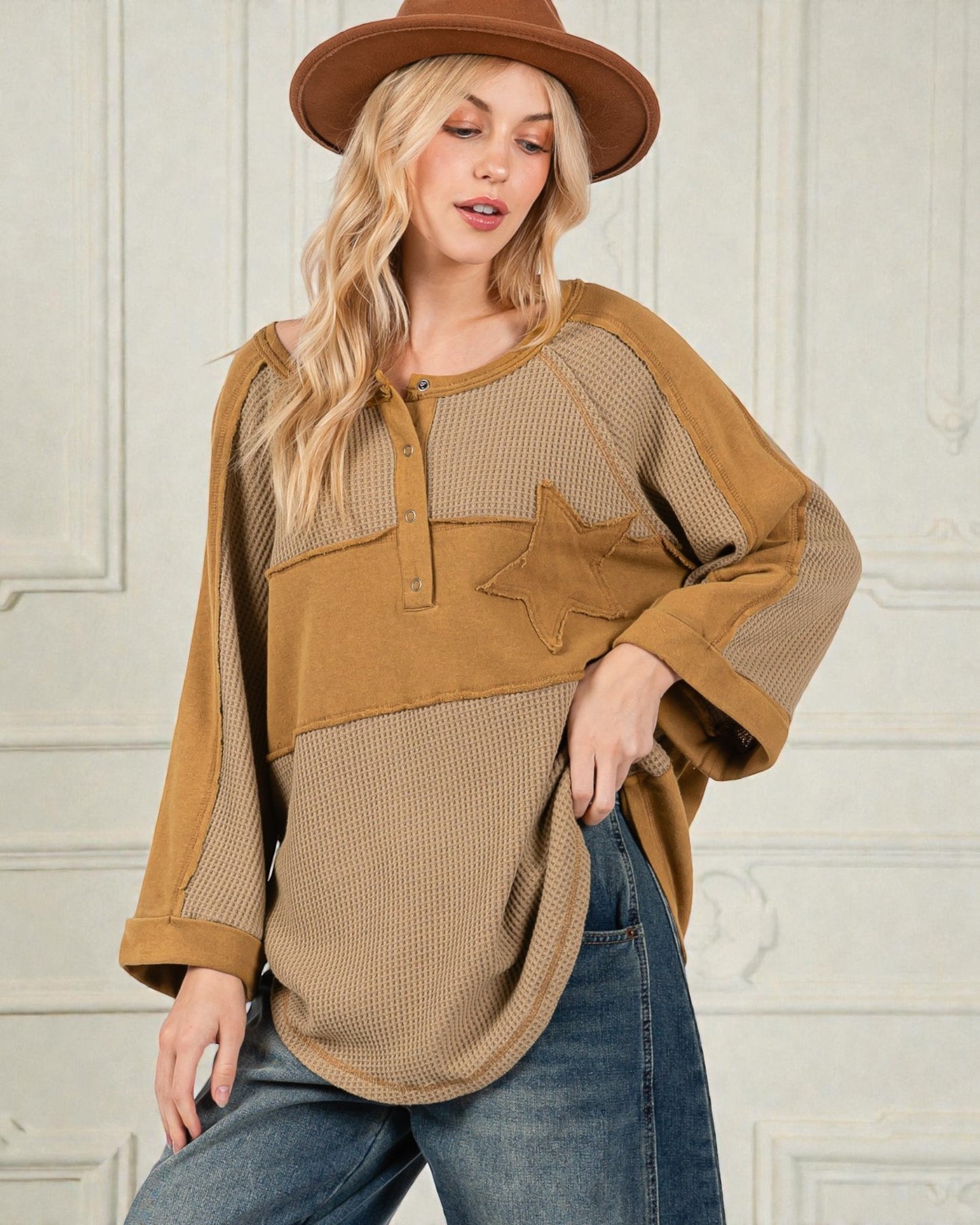 Woman wearing a brown and beige long-sleeve top with a star design, paired with blue jeans and a brown hat.