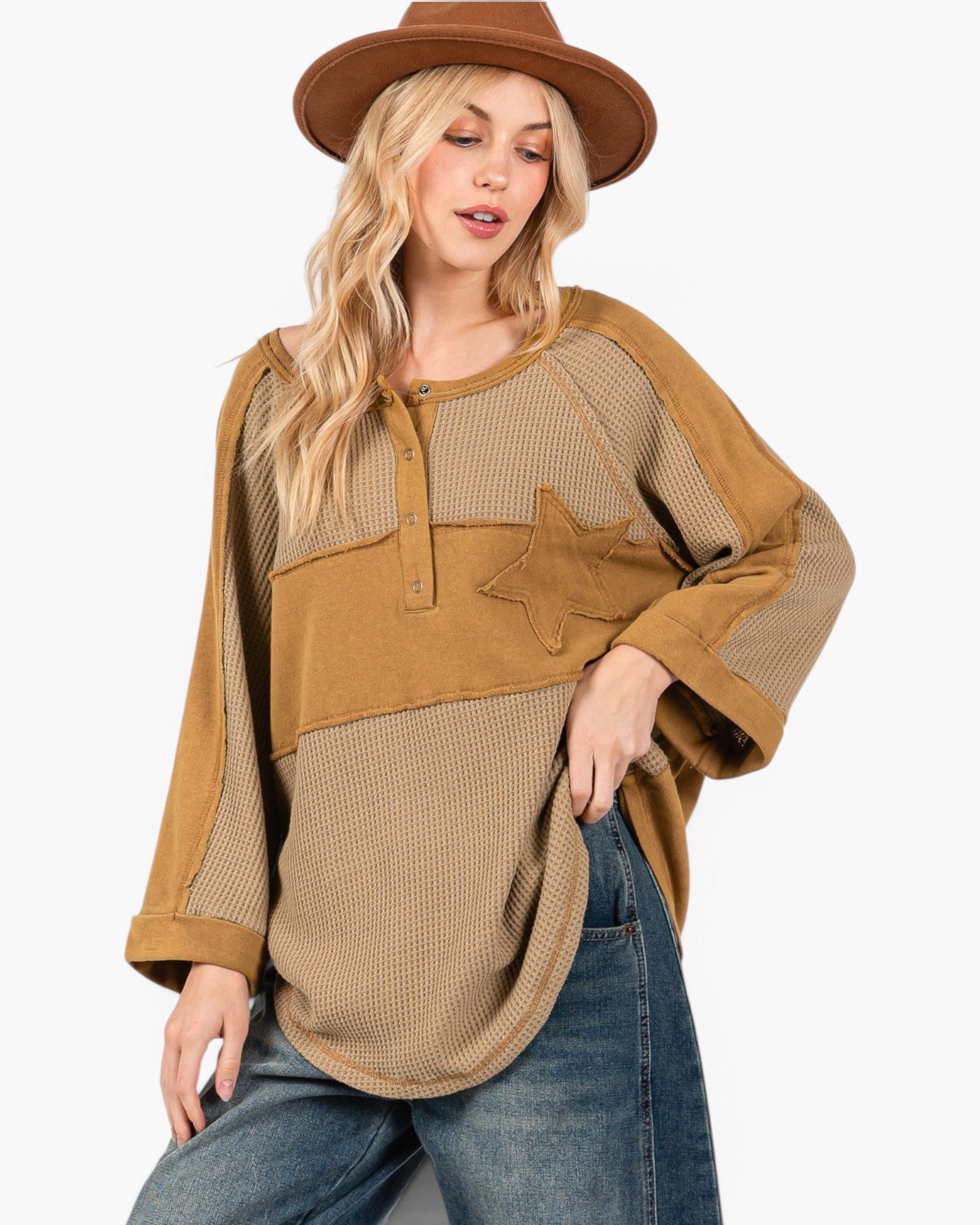A woman wearing a brown wide-brim hat and a tan and beige patchwork long-sleeve top with a star detail, paired with blue jeans, against a plain white background.