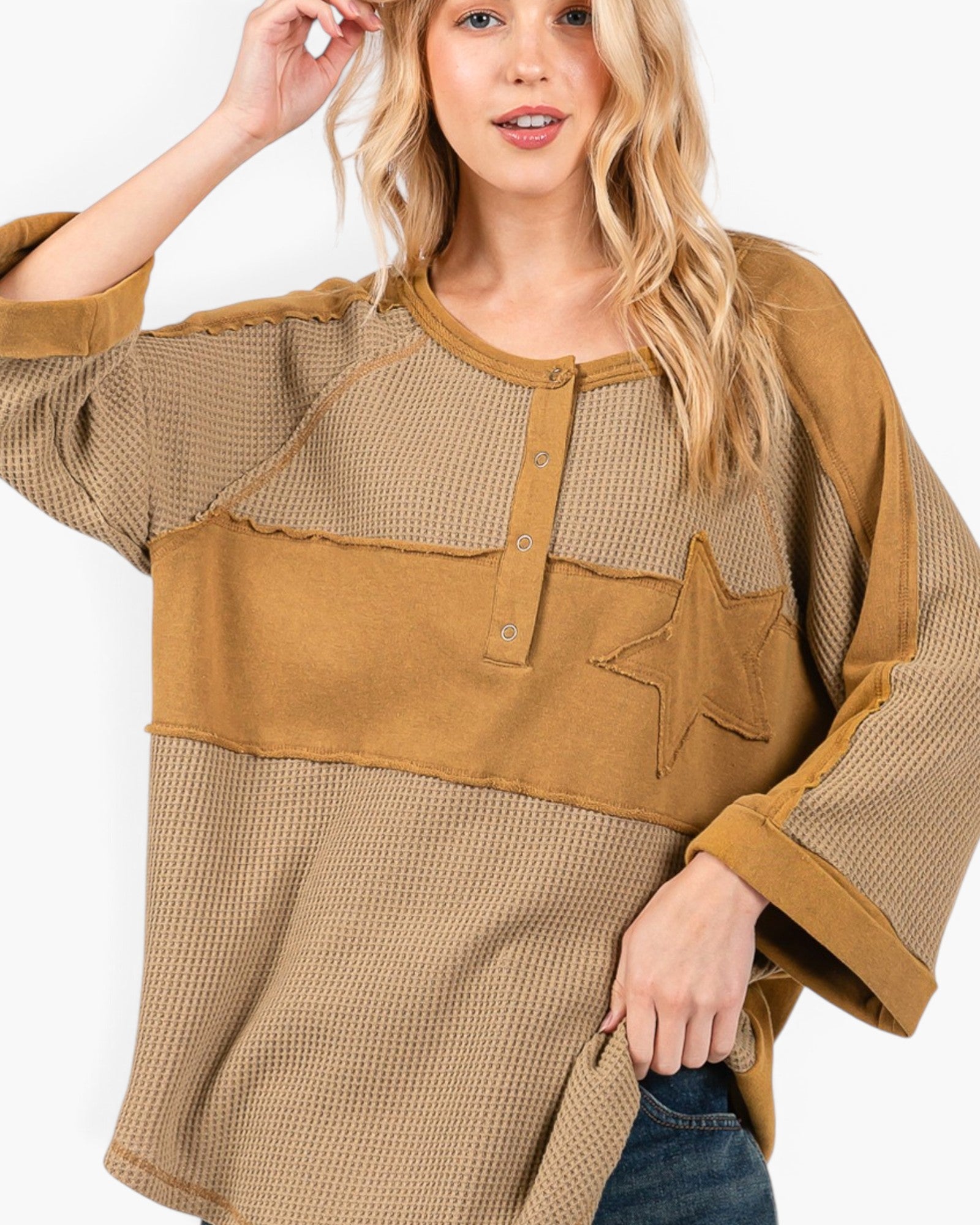 A woman wearing a loose, patchwork-style top with waffle-knit and solid fabric panels in tan and brown tones, featuring a star applique on the chest and a button placket.