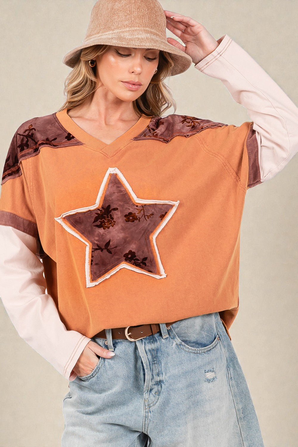 Person wearing an orange sweater with a star design, beige hat, and blue jeans on a neutral background