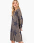 A woman modeling a long-sleeve, crewneck knit dress with a faded charcoal gray base and large dark navy star patterns, set against a plain white studio background.