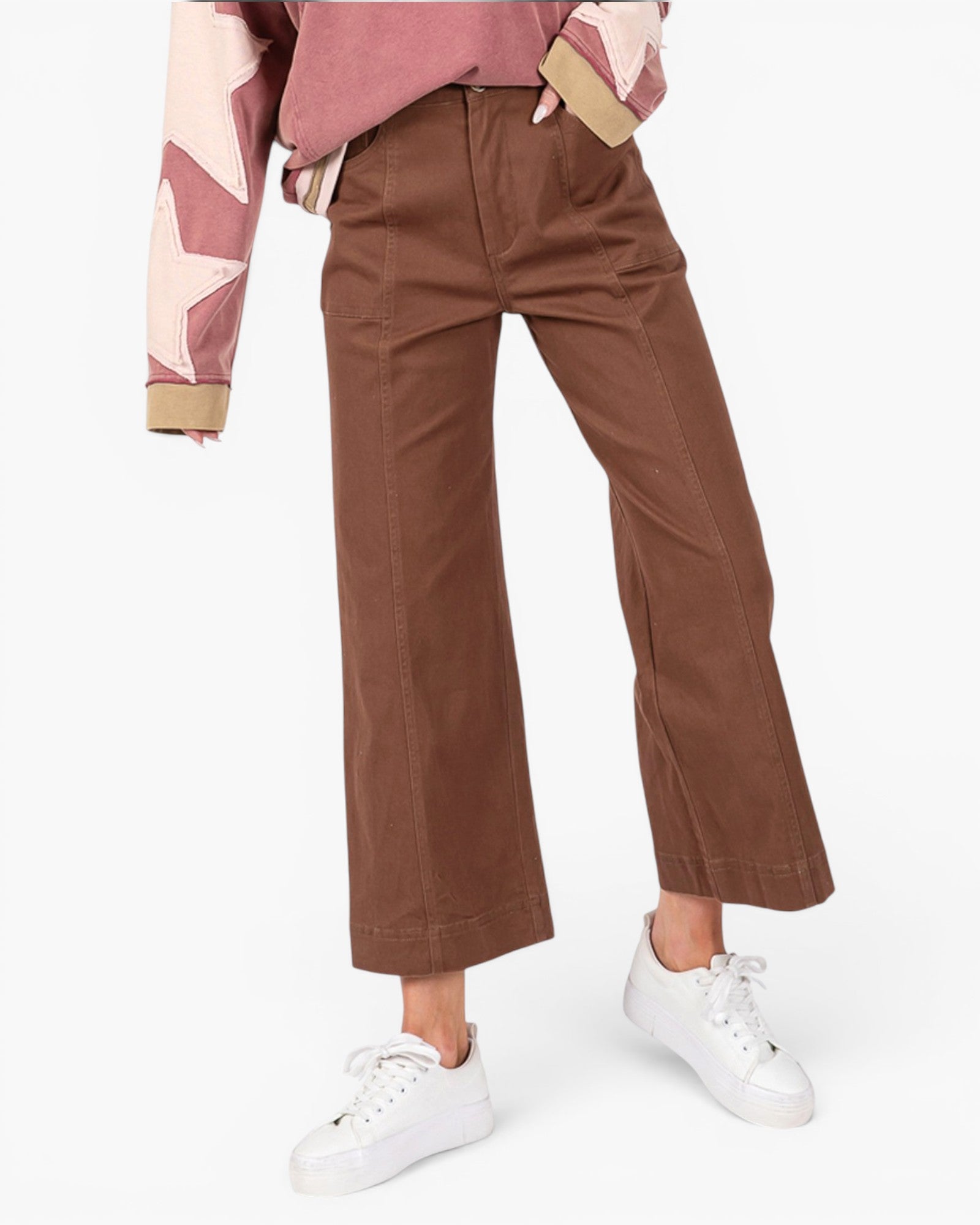 A person wearing brown wide-leg cropped pants with a pink patterned top and white sneakers, shown from the waist down on a light gray studio background.
