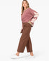 A woman modeling a dusty rose long-sleeve knit top with light pink geometric patchwork details on the sleeves, paired with brown cropped trousers and white sneakers, against a plain white studio background.