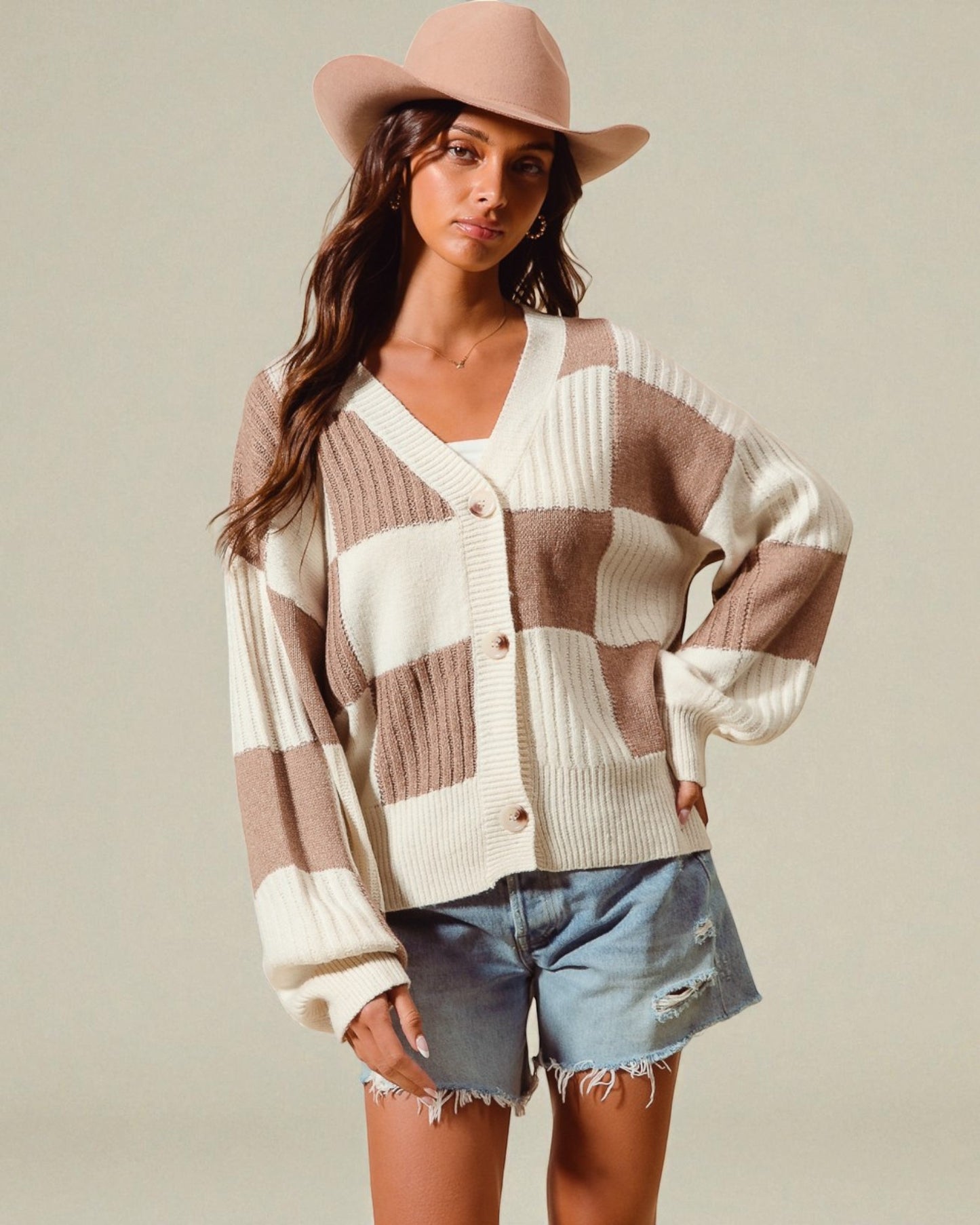 Woman wearing a checkered cardigan, denim shorts, and a pink hat on a plain background