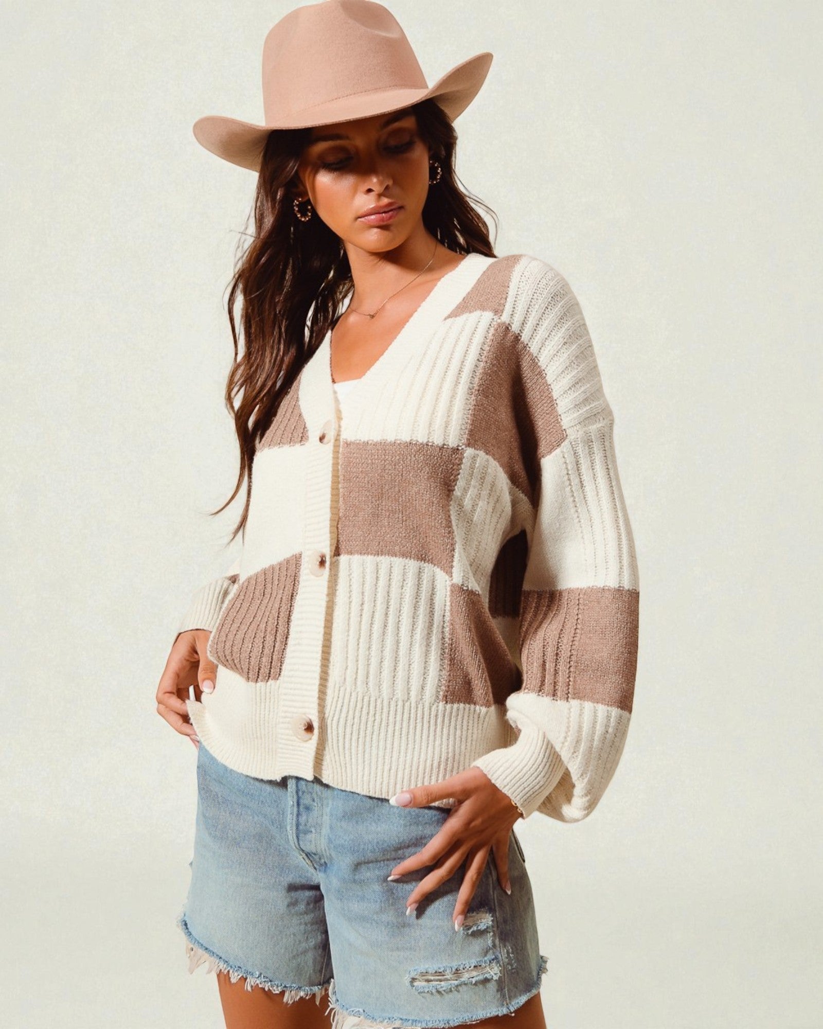 Woman wearing a patchwork sweater and denim shorts with a beige hat on a light background