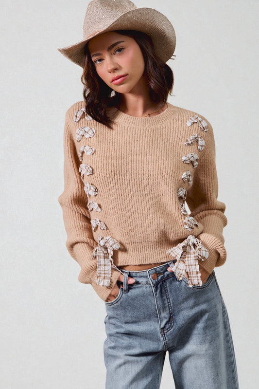 Woman wearing a beige sweater with floral details, blue jeans, and a beige hat on a white background
