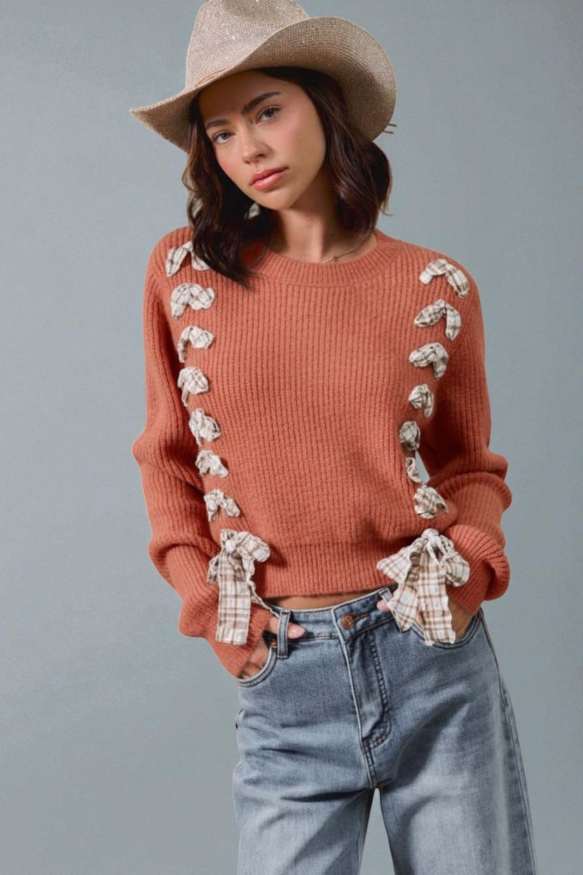Woman wearing a rust-colored sweater with floral details, blue jeans, and a beige hat against a gray background