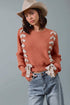 Woman wearing a rust-colored sweater with floral details, blue jeans, and a beige hat against a gray background