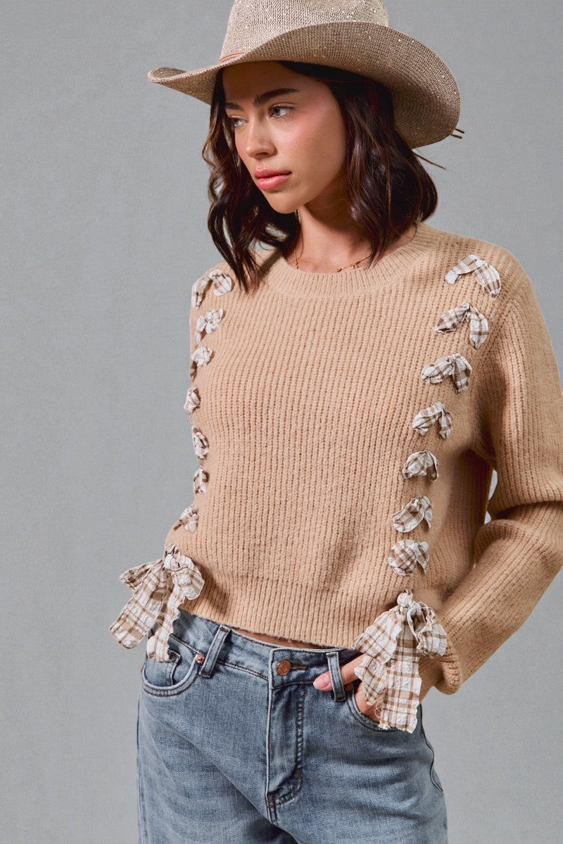 Woman wearing a beige sweater with ruffled details, blue jeans, and a beige hat against a gray background