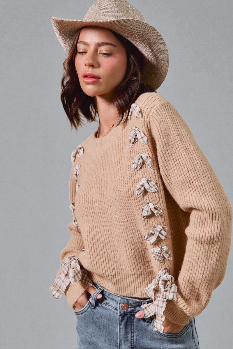 Woman wearing a beige sweater with bow details, denim jeans, and a beige hat on a gray background