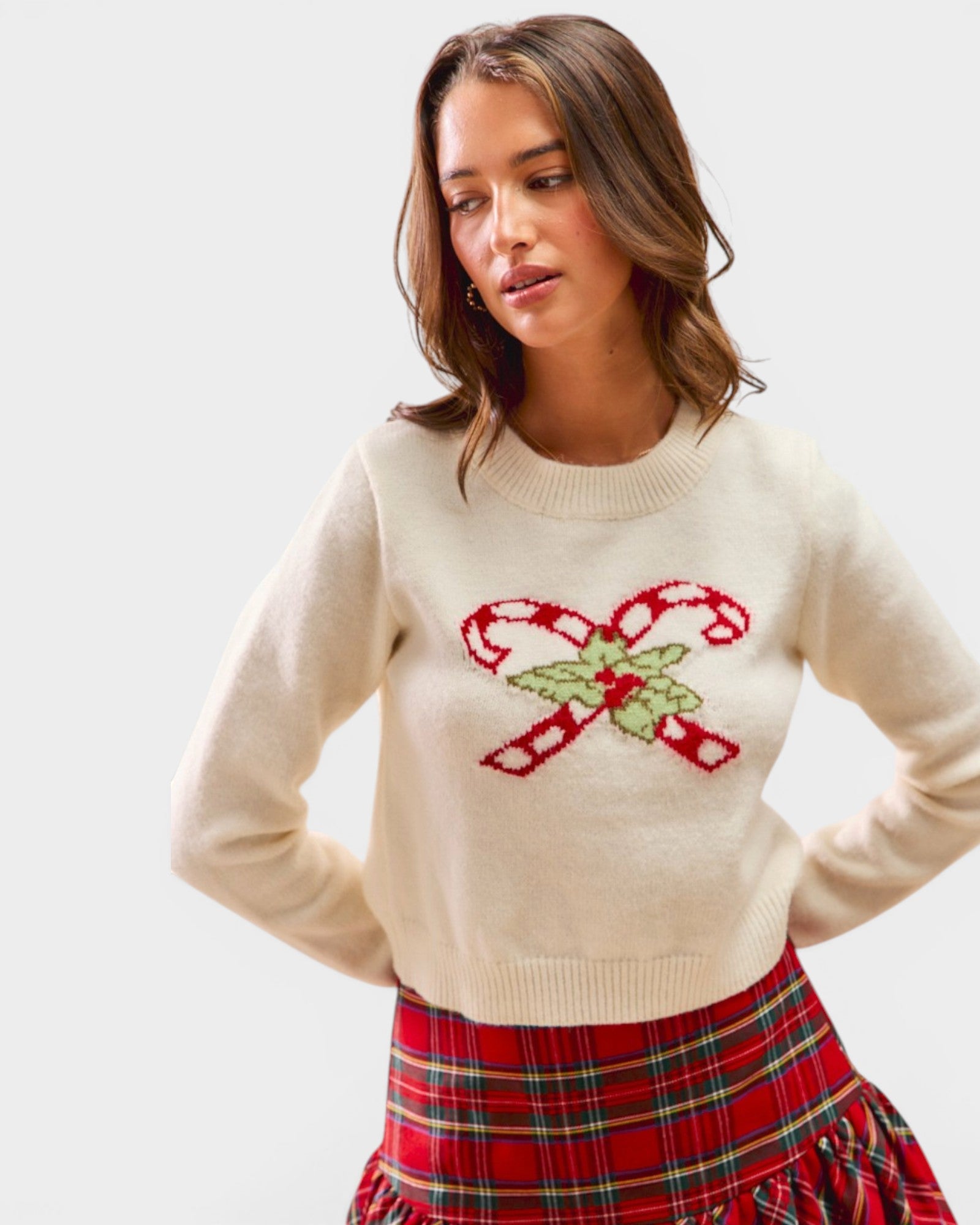 Woman wearing a cream sweater with candy cane design and plaid skirt on a white background