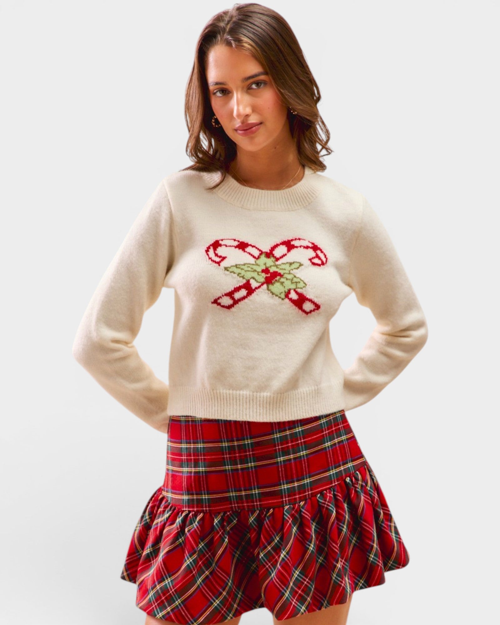 Woman wearing a cream sweater with candy cane design and a red plaid skirt on a white background