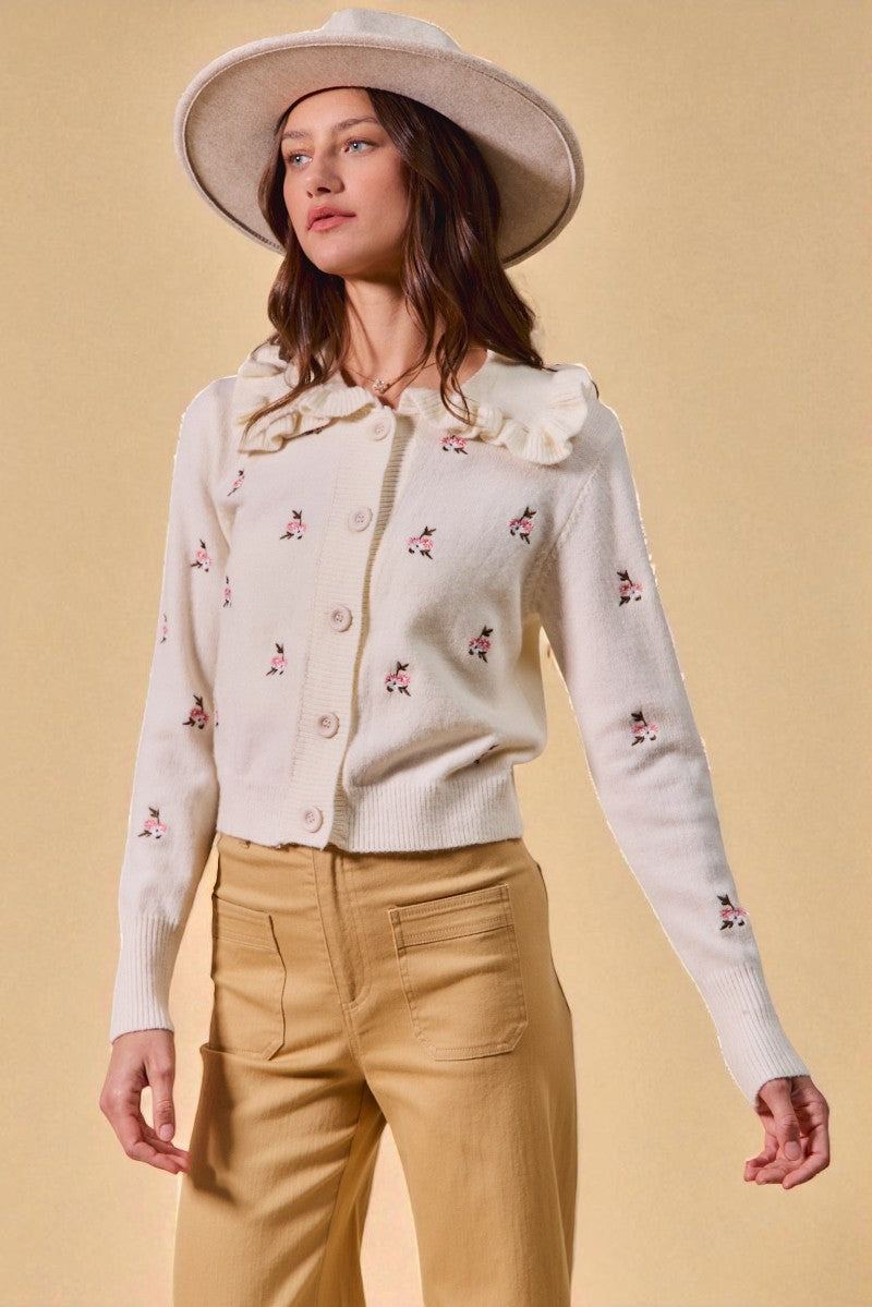 Woman wearing a white embroidered cardigan and beige pants against a beige background