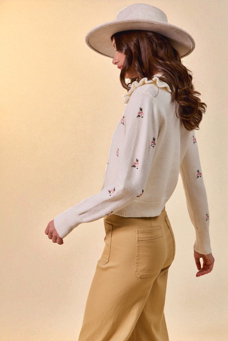 Woman wearing a white floral cardigan and beige pants with a wide-brimmed hat on a beige background