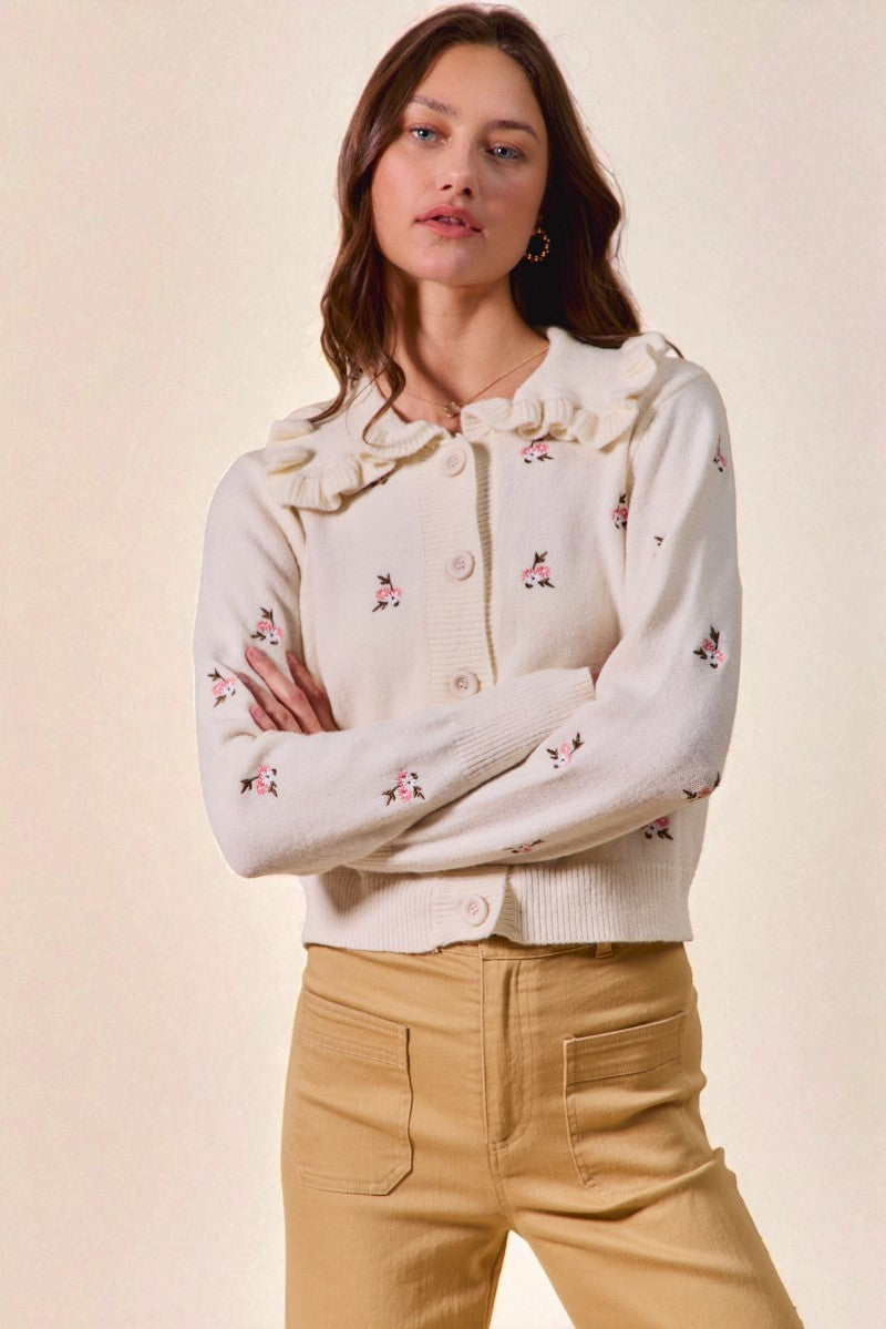 Woman wearing a white embroidered cardigan with beige pants on a plain background