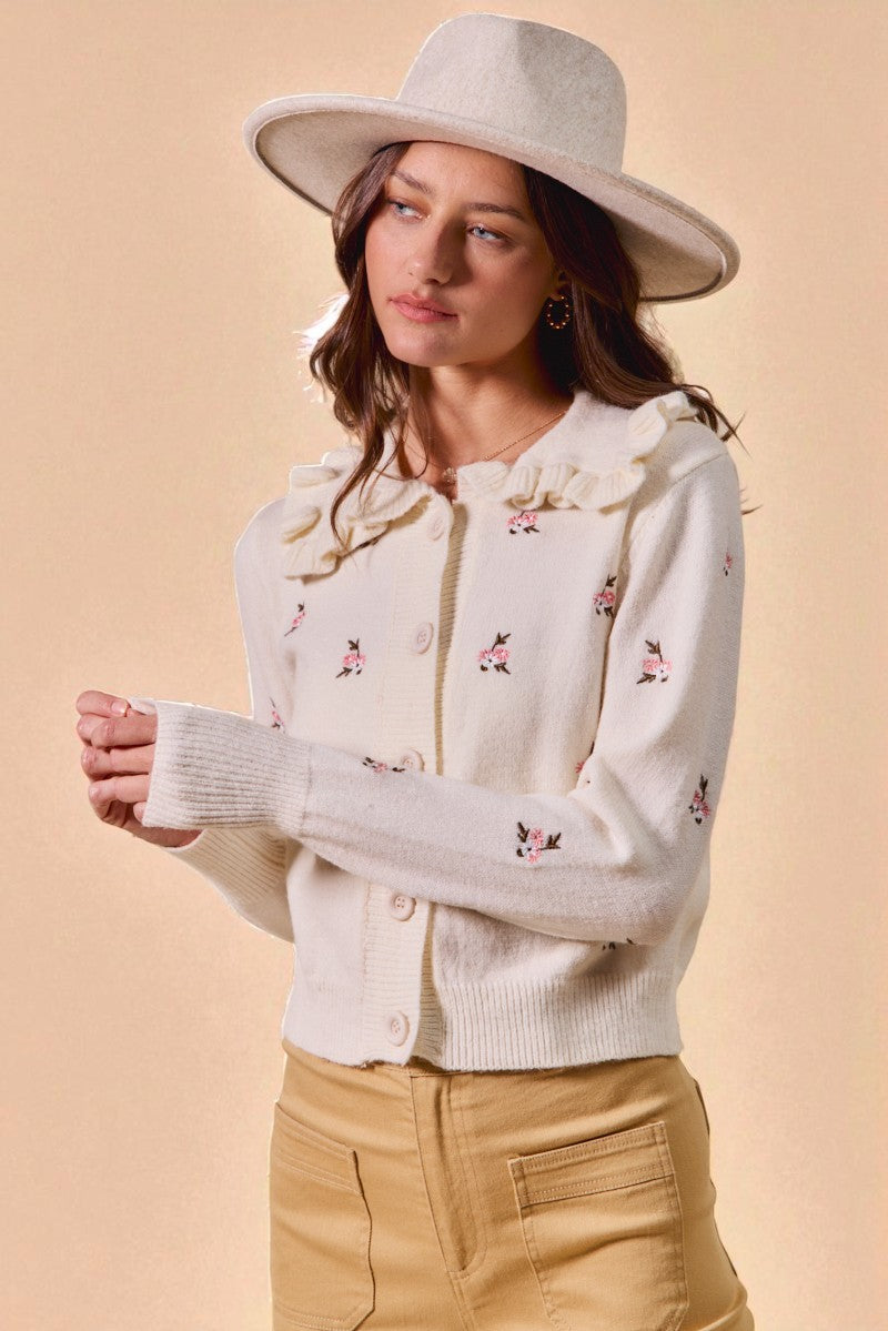 Woman wearing a white floral embroidered cardigan and beige hat against a beige background